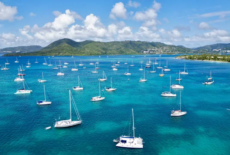Aerial of a Caribbean anchorage with a catamaran in the foreground and many sailboats off a green coast