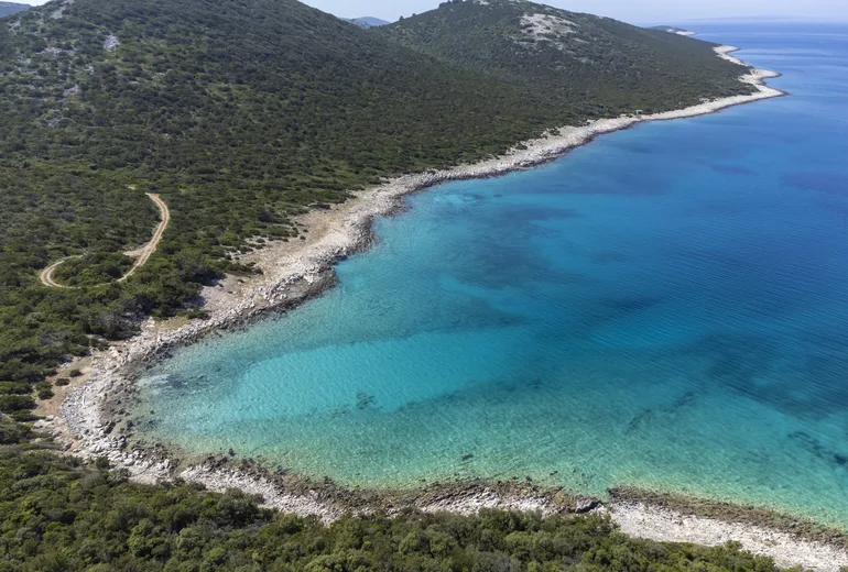Aerial of a remote turquoise cove with a white pebble rim and scrub hills in Croatia
