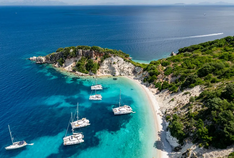 Aerial of charter yachts anchored off a white pebble cove on a green Ionian islet, Greece
