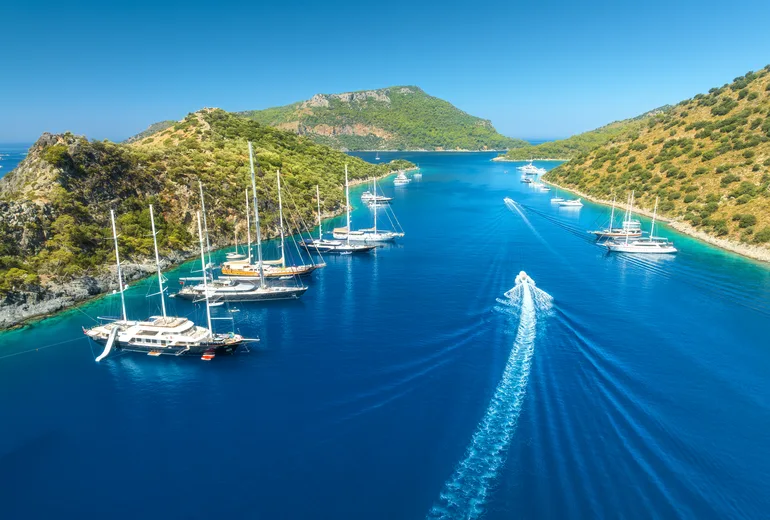 Aerial of gulets and yachts anchored in a narrow turquoise channel between pine hills, Turkiye