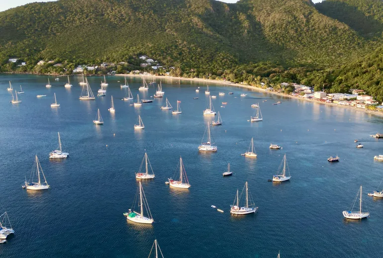 Aerial of monohull sailboats anchored off a beach village in evening light, green mountains behind