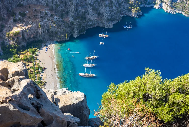 Gulets anchored in the deep blue Butterfly Valley cove beneath sheer cliffs and a beach, Türkiye