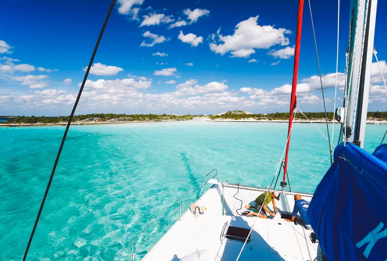 Sailboat foredeck with two crew approaching a low sandy cay over turquoise banks