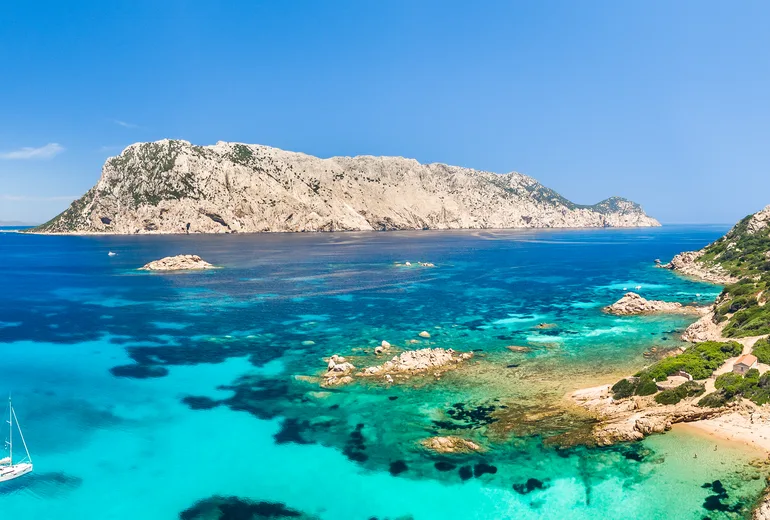 Tavolara island limestone massif over turquoise shallows with an anchored sailboat, Sardinia