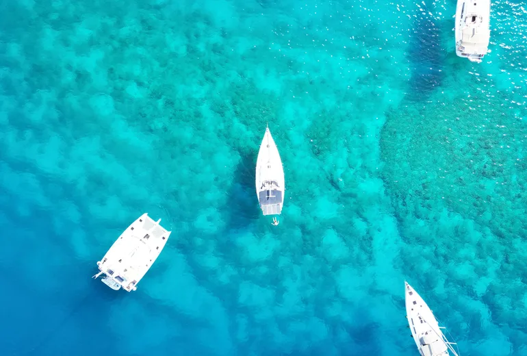Top-down aerial of a catamaran and sailboats anchored over turquoise reef shallows