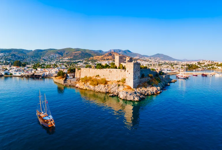 Wooden gulet sailing past Bodrum castle walls with the town and hills behind, Türkiye